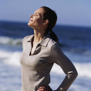 menopause_relaxation-bien-etre-serenite-nature Femme détendue au bord de la mer, les yeux fermés, illustrant relaxation, sérénité et bien-être naturel.